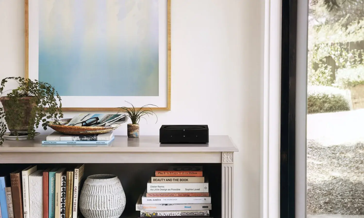 A Sonos Amp on a white shelf beside books and a plant, doors open to a garden