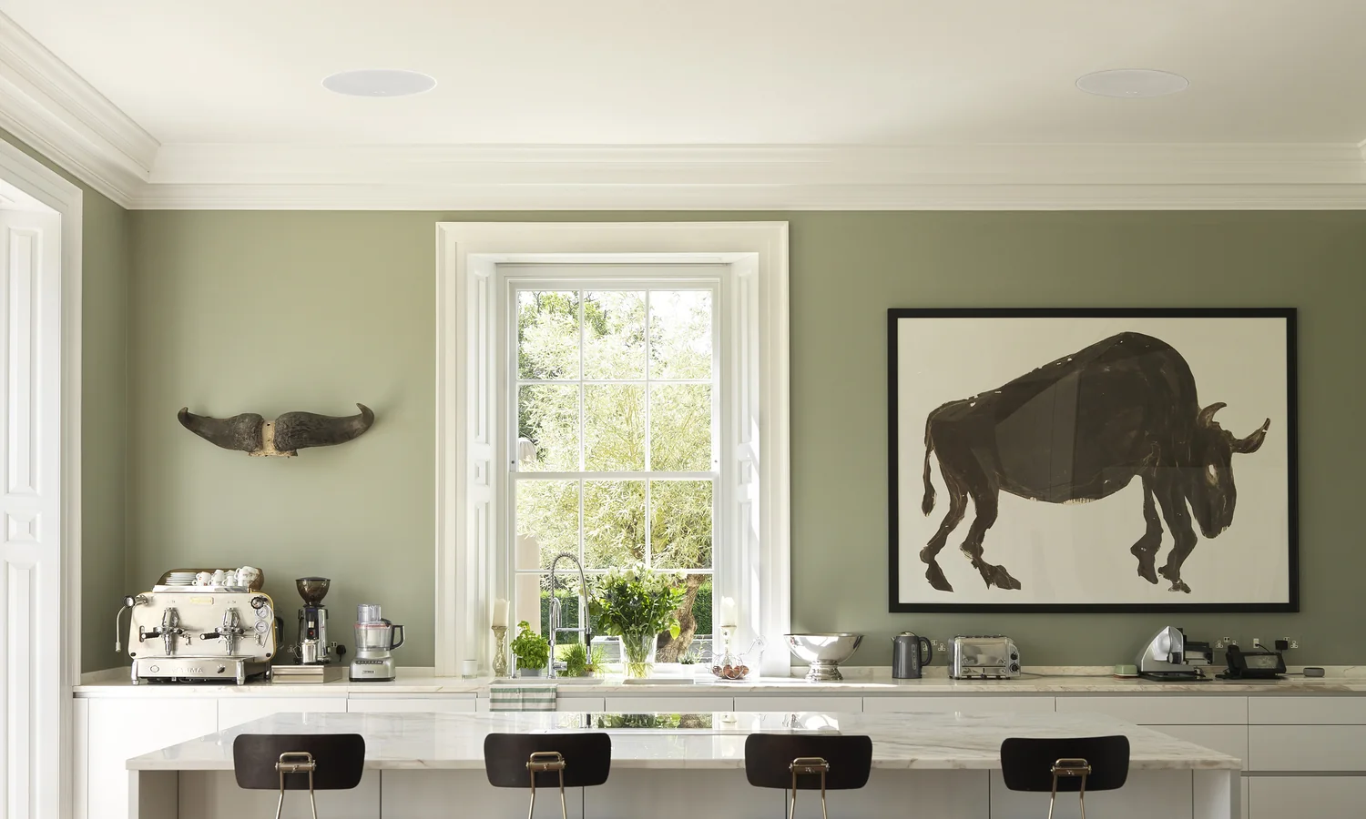 A sage-green kitchen with two KEF in-ceiling speakers flush in the plaster above the island