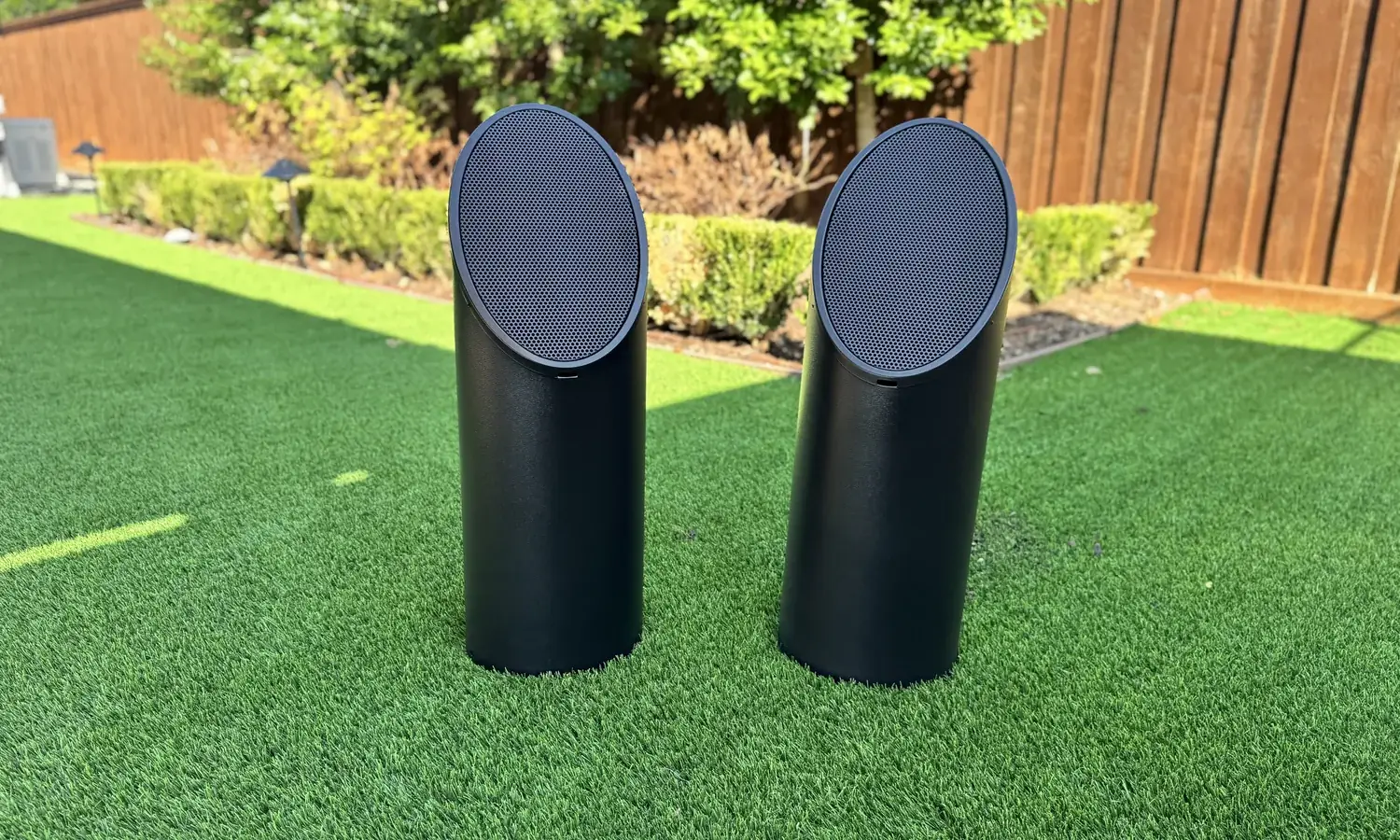 A pair of bollard speakers standing on turf with a hedge and fence behind