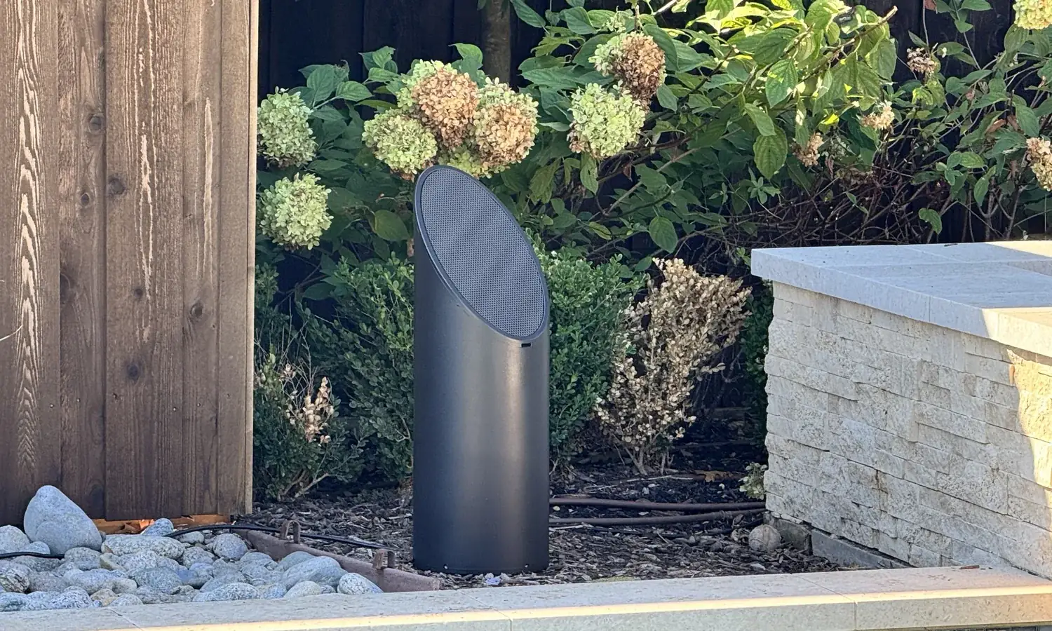 A single bollard speaker in a planting bed between a cedar fence and a stone wall