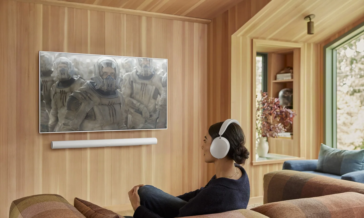 A woman in Sonos Ace headphones watching television in a cedar-paneled room with an Arc soundbar under the set