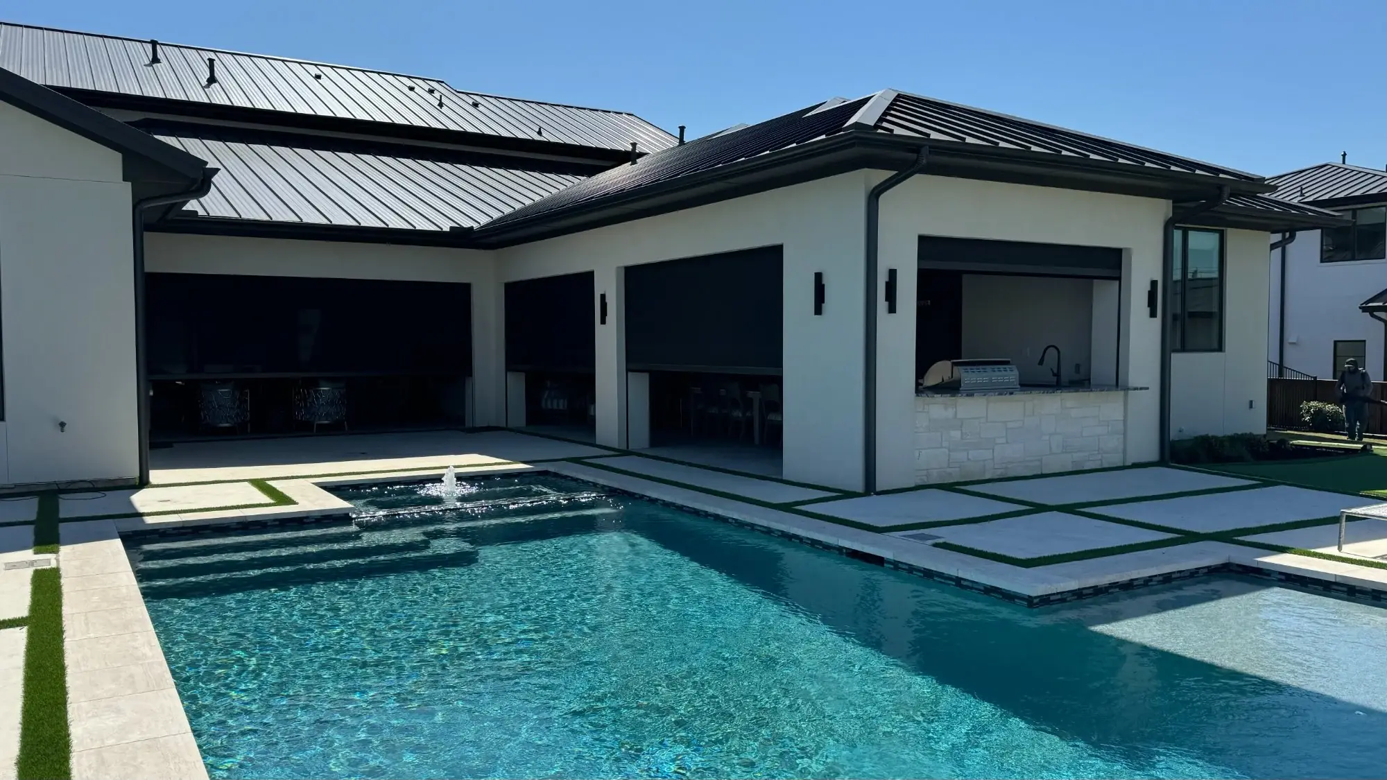A modern pool cabana with dark exterior shades lowered across its open sides, pool and fountain in the foreground