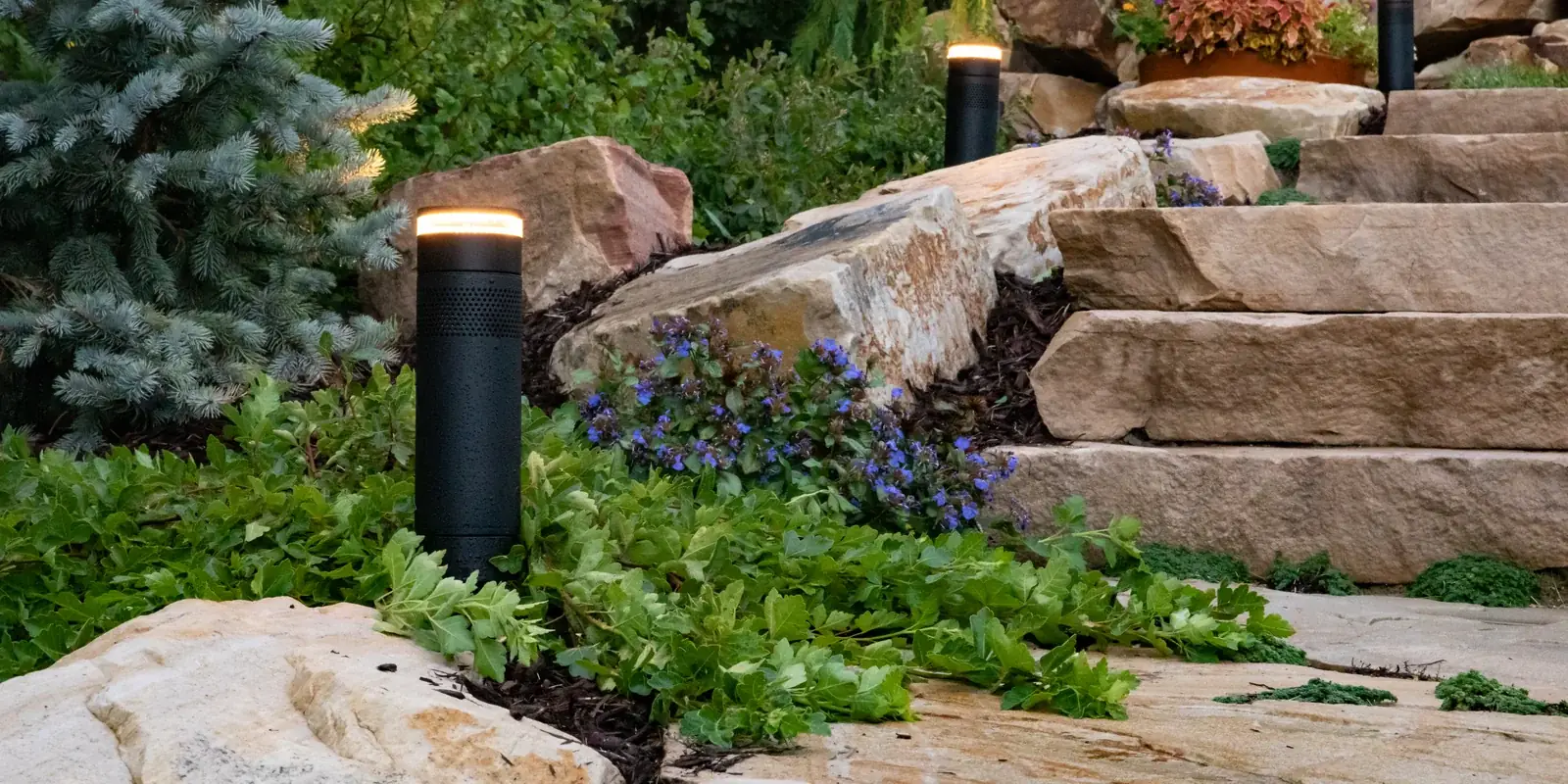 Bollard speakers with lit rings along a stone stair at dusk, planting on both sides
